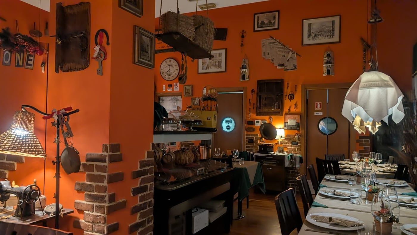 La sala dell'Osteria al Portone: pareti arancioni, mattoni a vista, oggetti d'antiquariato appesi e tavoli apparecchiati con tovaglioli verdi