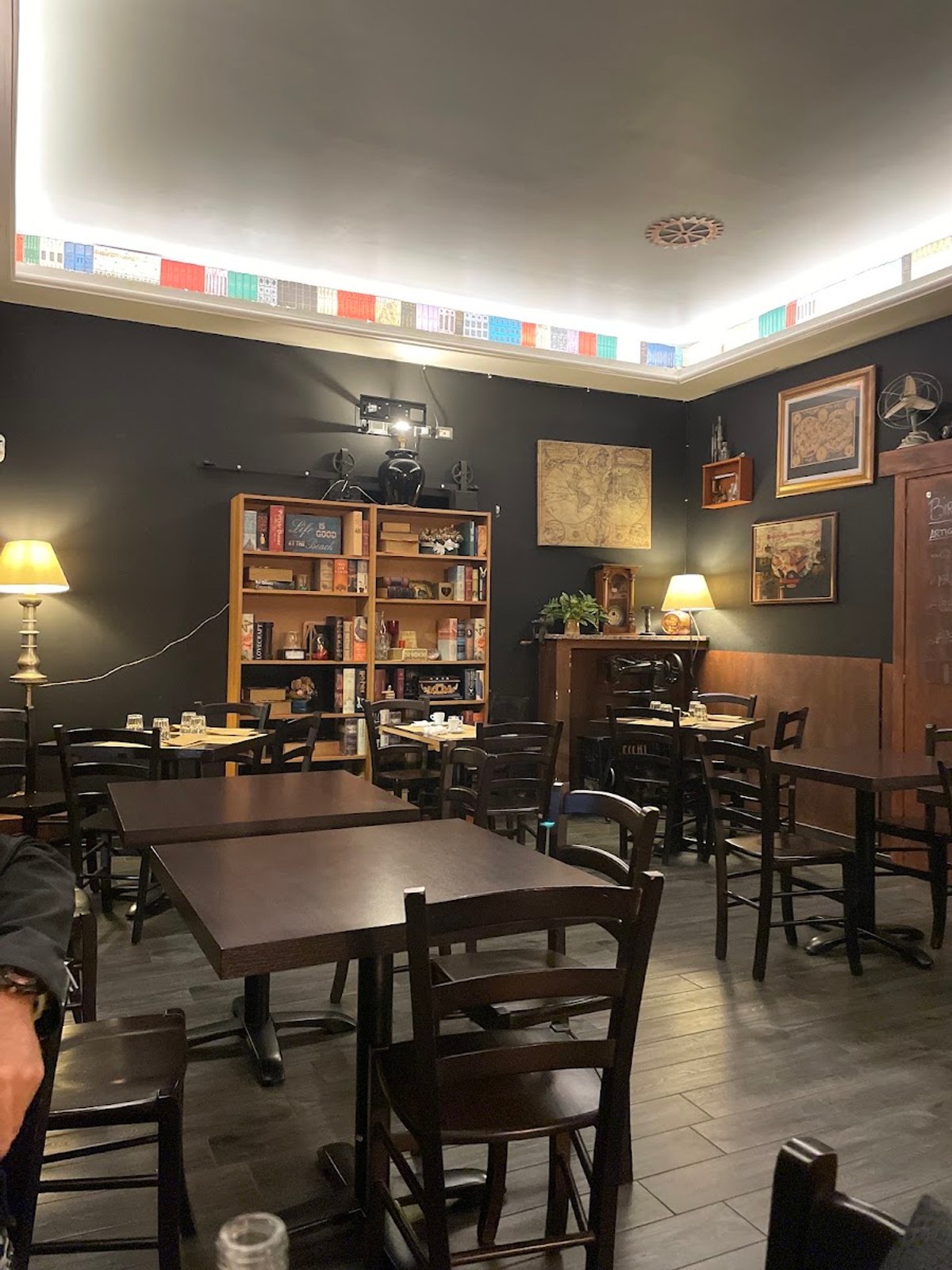 Una sala con la libreria piena di libri, mappe antiche appese e lampade da tavolo accese