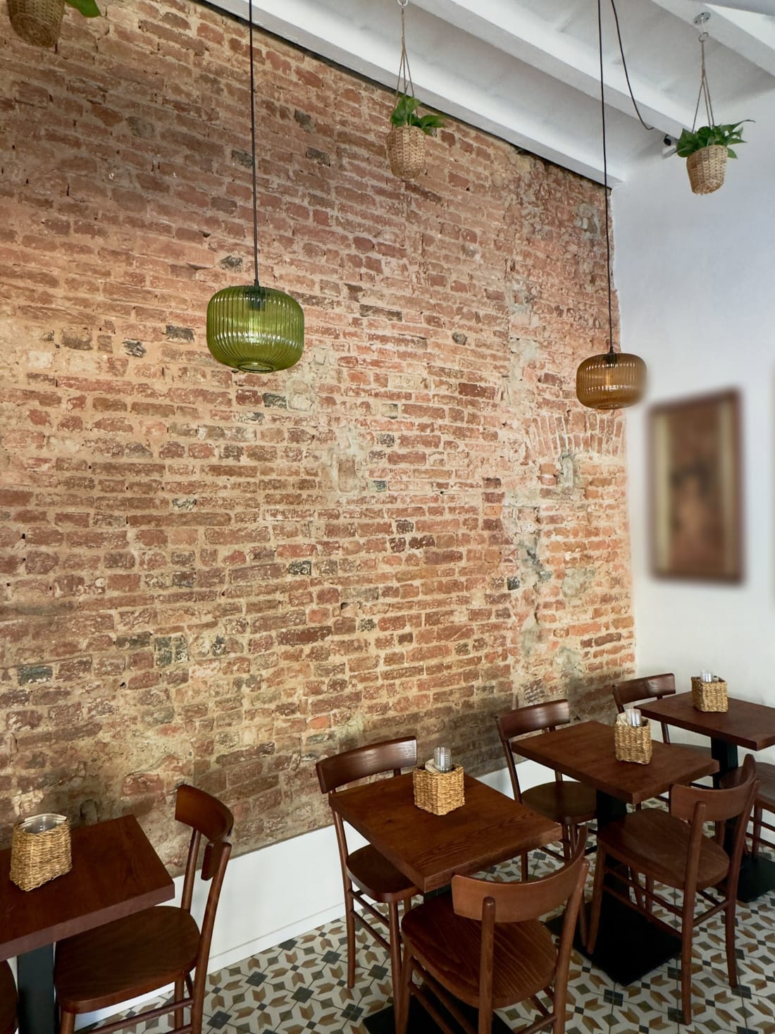 La sala nuova: il muro di mattoni a vista, le lampade di vetro rigato verde e ambra, le piante nei cestini, i tavoli di legno scuro e le cementine.