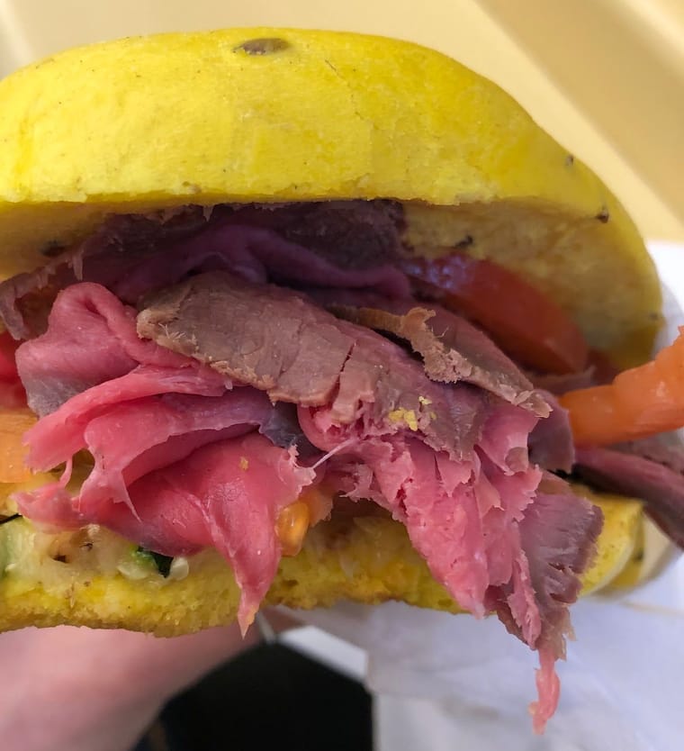 Un panino giallo alla curcuma col roast beef, le zucchine grigliate e il pomodoro, tenuto in mano.