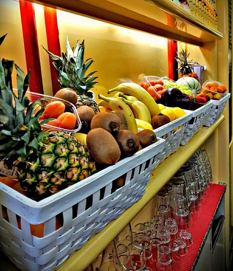 I cesti di frutta sul banco: ananas, kiwi, banane, arance, davanti alle mensole gialle coi filetti rossi.