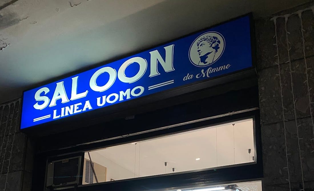 L'insegna blu illuminata di sera, «Saloon Linea Uomo da Mimmo» col loro logo, sopra la vetrina
