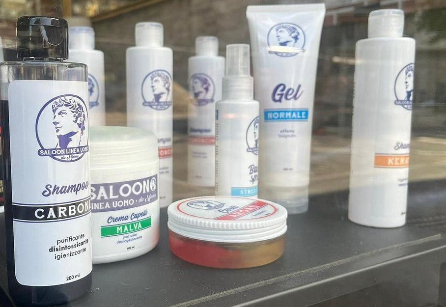Flaconi e vasetti bianchi con il loro logo, la testa classica nel cerchio blu: shampoo al carbone, crema capelli alla malva, gel, cera