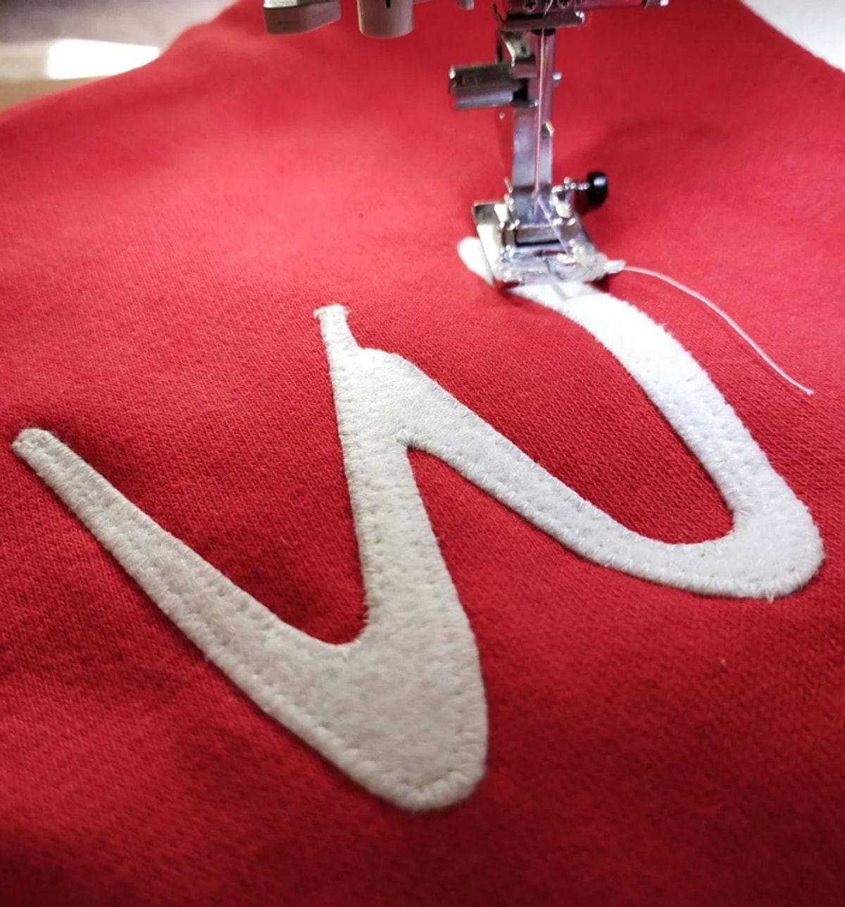 Una W di panno cucita a mano su una maglia rossa, con il piedino della macchina da cucire e il filo ancora attaccato