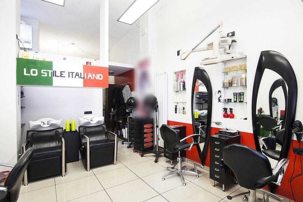 Il salone vuoto: pareti bianche, due lavatesta neri sotto la scritta LO STILE ITALIANO sui colori della bandiera, le poltrone davanti agli specchi ovali neri, una fascia rossa lungo il muro.