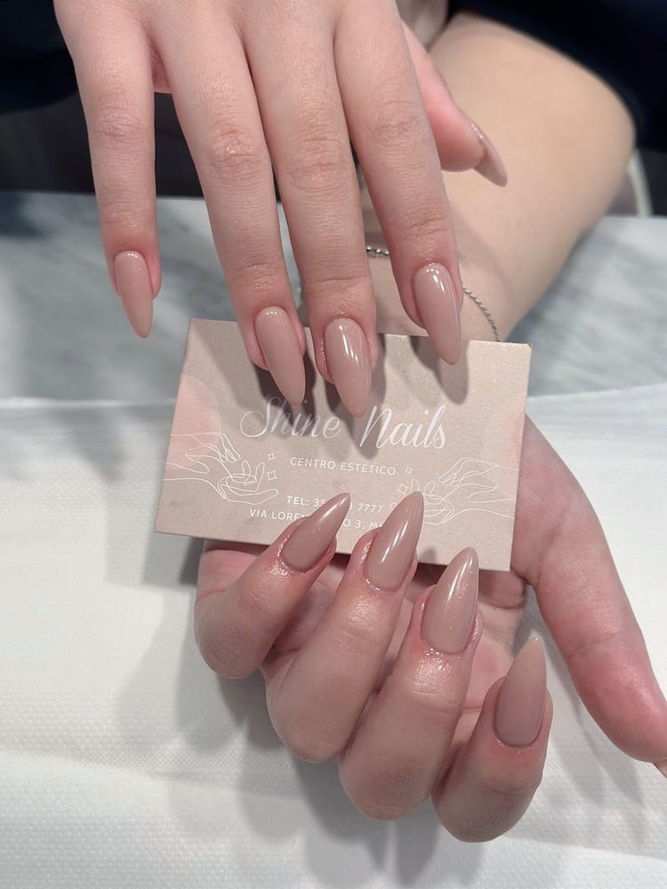 Unghie lunghe a mandorla color nude, e il biglietto rosa di Shine Nails fra le dita.