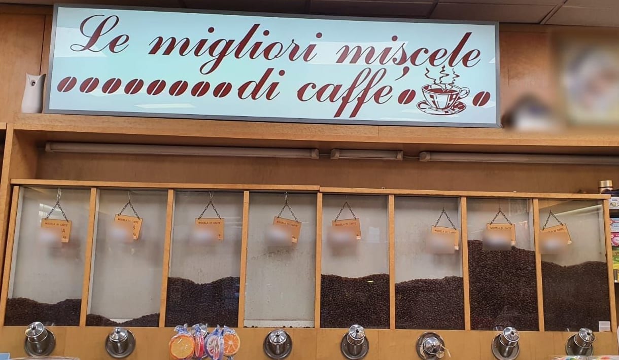 L'insegna azzurra «Le migliori miscele di caffè» in corsivo bordeaux, con i chicchi al posto delle o, sopra la parete degli otto silos di vetro pieni di chicchi, coi cartellini e i dosatori d'acciaio.