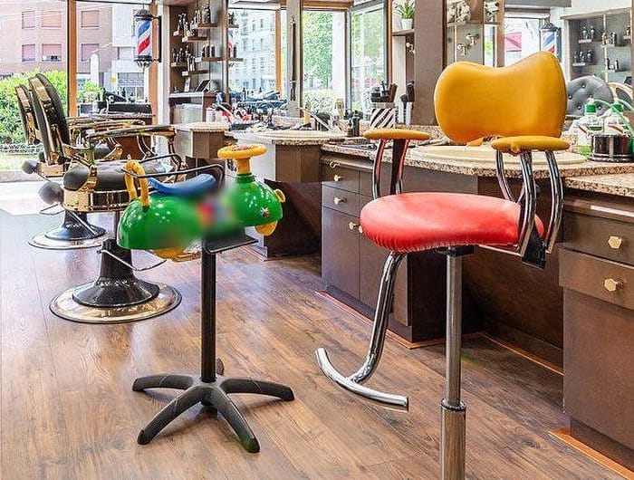La poltroncina per bambini a forma di macchinina verde col volante giallo, accanto uno sgabello giallo e rosso e, dietro, le poltrone e il palo da barbiere