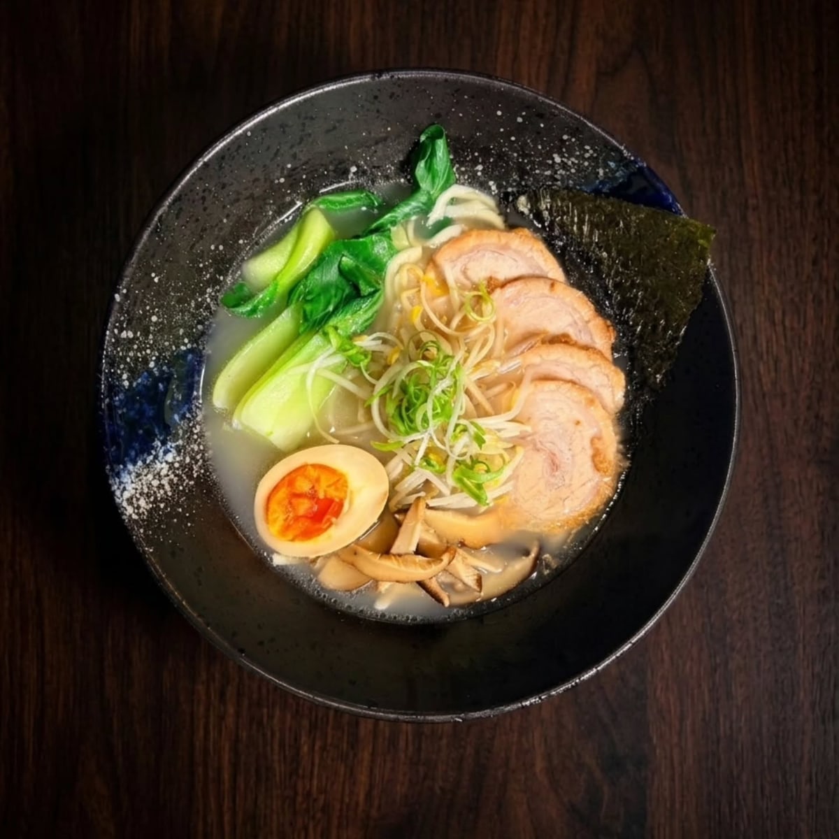Una scodella di ramen tonkotsu vista dall'alto: brodo chiaro, fette di maiale chashu, uovo marinato, pak choi, germogli, funghi shiitake e alga nori